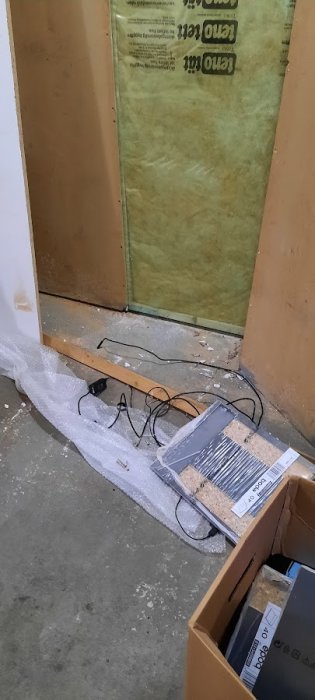 Delvis isolerad vägg med mineralull och synliga träreglar, kabel och öppen kartong med byggmaterial på golvet.