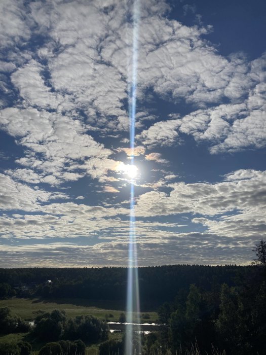 Sol som skiner genom moln över ett landskap med träd och ett litet vattendrag.