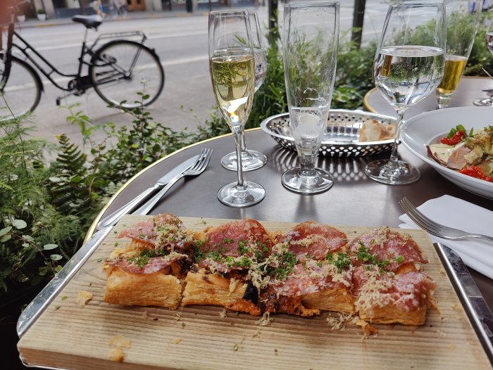 Fransk pizza med skivad salami på en träbräda, två glas vin och ostron i bakgrunden, utomhusmiljö med cykel.