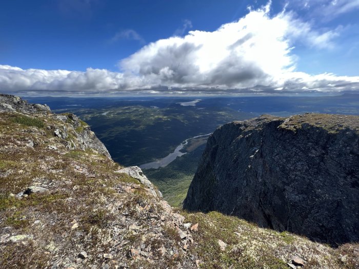 Utsikt från bergstopp med klippkanter, lummig dal och en flod under delvis molnig himmel.