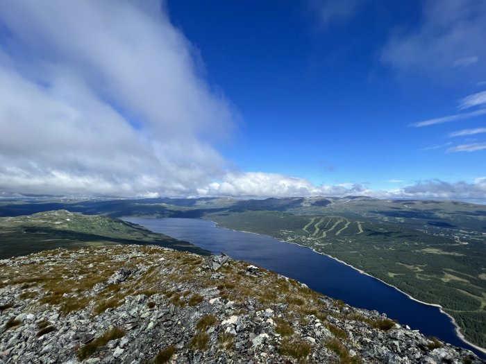 Utsikt från bergstopp över sjö och skog med delvis molntäckt himmel.