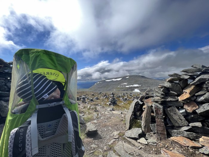 Bebis i bärsjal med myggnät framför stenformation med bergslandskap och molnhimmel i bakgrunden.