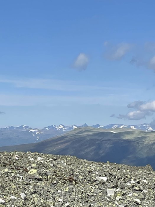 Vy från ett stenigt berg mot en horisont med snöklädda bergstoppar under en klarblå himmel.