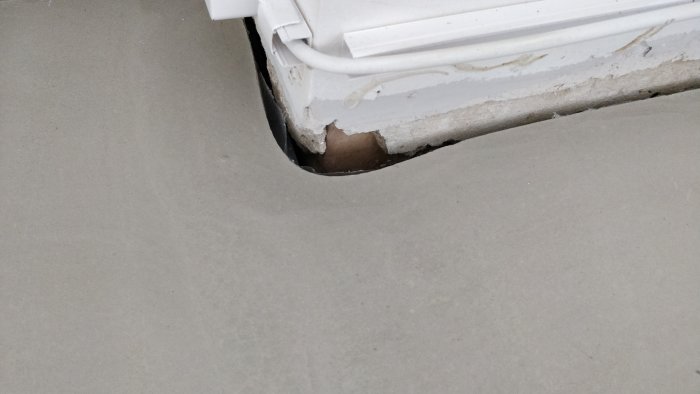 Skadad del av betonggolv och vägg nära dörrfoder och elledningar.