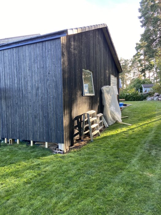 Svartmålad träfasad på hus med gräsmatta, staplade stolar och en täckt objekt på sidan.