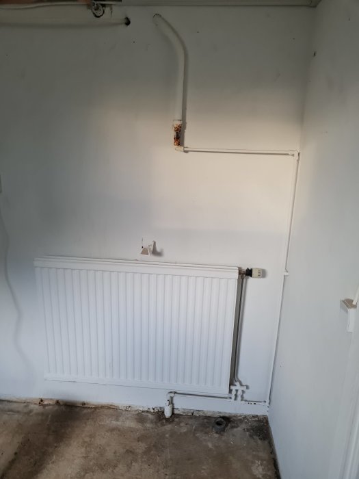 Vit vägg med radiator och synliga rör, inklusive ett rostigt rör, ovanför betonggolv.