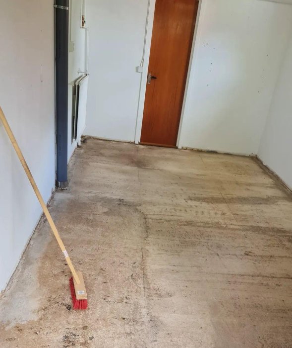 Ett rum med betonggolv under renovering, sopkvast på golvet vid öppen dörr.