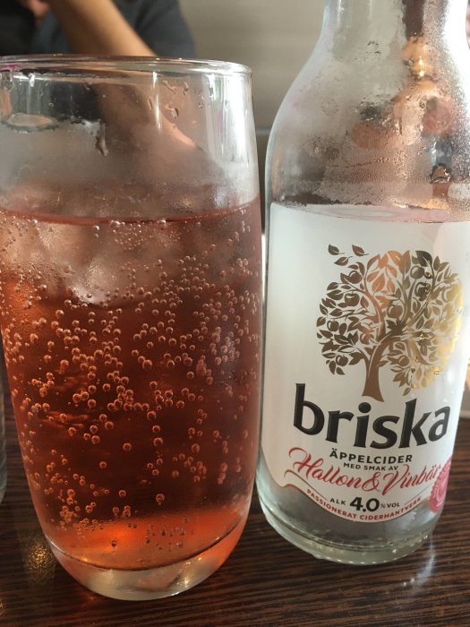 Ett glas och en flaska hallon- och vinbärsäppelcider från Briska på ett bord.