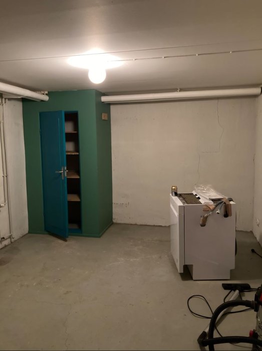 Källarrum före renovering med öppen grön dörr, skåp och betonggolv, synliga rör i taket.