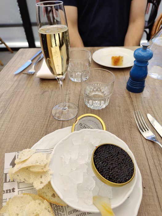 Ett glas champagne, en burk med kaviar på is, brödbitar och en tom tallrik på ett restaurangbord.