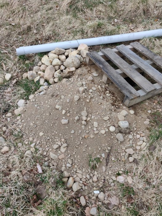 Stenig mark med grus och större stenar bredvid en träpall och ett vitt plastavrinningsrör.