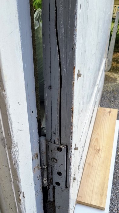 Närbild på en vit dörr med en lagad spricka vid gångjärnets infästning, vagnsbultar och spikar synliga.