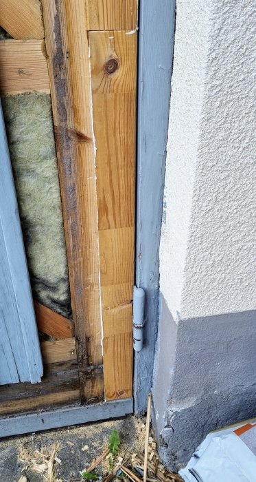 Reparerat trä på dörrkarm med nytt trä och synligt gångjärn mot grå husvägg.