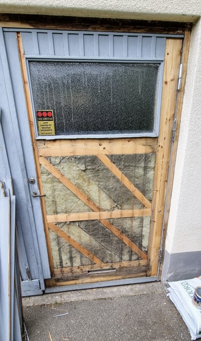 Renoveringsarbete på en dörr, med plywood monterad över isolering, markerade reglar och delvis täckt fönster.