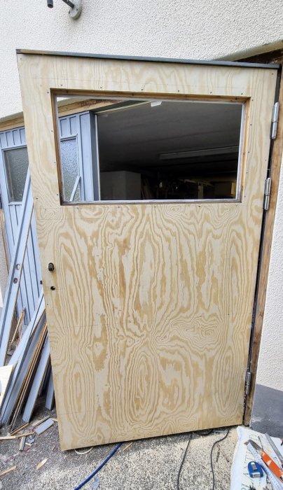 Dörr klädd med ny 4mm plywood, urfräst fönsteröppning, och verktyg synliga på marken.