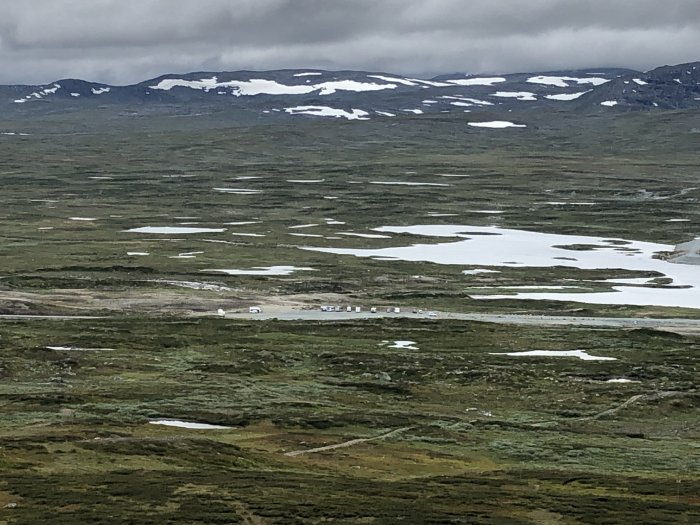 Utsikt över fjällandskap med smältande snö och gröna områden med synlig väg och byggnader.