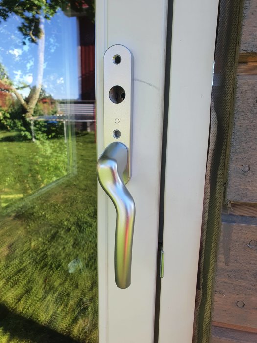 Fönsterhandtag och låsmekanism på vit fönsterdörr i förgrunden med trädgårdsutsikt speglad i glaset.