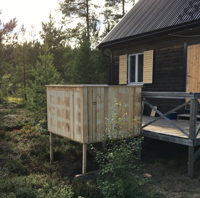 Nybyggt, obehandlat träutbyggnad till en mörk stuga med veranda och skogsbakgrund.