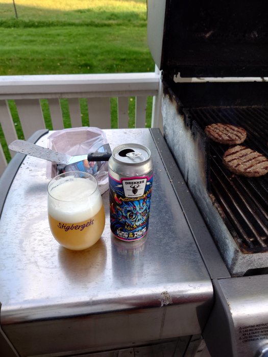 Ölburk av märket Drekker och ett ölglas på en grill med hamburgare i utomhusmiljö.