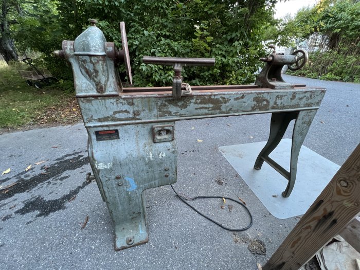 En äldre, begagnad svarv med synliga slitage och rost, står på en asfalterad yta utomhus.