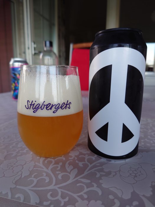 Ölglas märkt med "Stigbergets" fyllt med gyllene öl och en burk med peace-symbol vid sidan på ett bord.