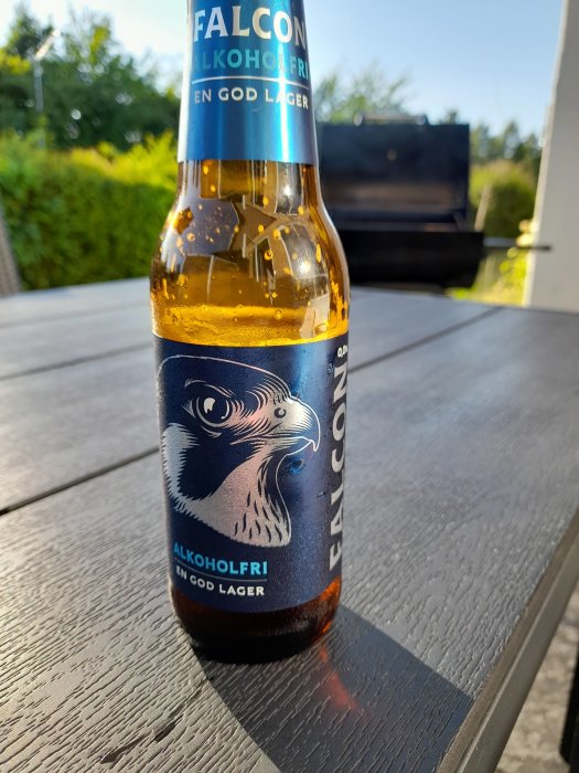 En ölflaska märkt Falcon Alkoholfri på ett bord utomhus med en grill i bakgrunden.