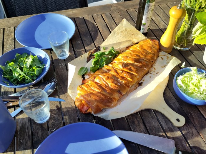 Hemgjord pizzafläta på bakplåtspapper bredvid sallad och tomatsåsflaska på ett träbord utomhus.