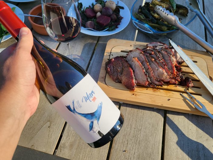 En hand håller en vinflaska med etiketten "Macia Batle" bredvid ett grillat måltid på en träterrass.