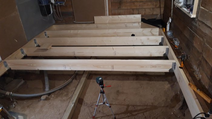 Pågående golvläggningsarbete med synliga träbjälkar och ett laserverktyg på ett stativ i en byggnad under renovering.
