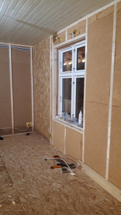 Pågående isoleringsarbete i rum med OSB-skivor på väggar, fönster och verktyg på golvet.