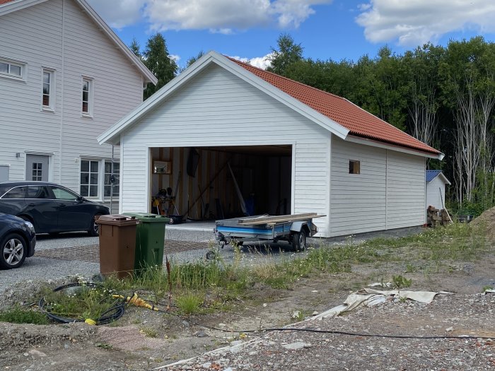 Nybyggt vitt garage med öppen port och rött tak intill en bostadsbyggnad, omgivet av grus och grönska.