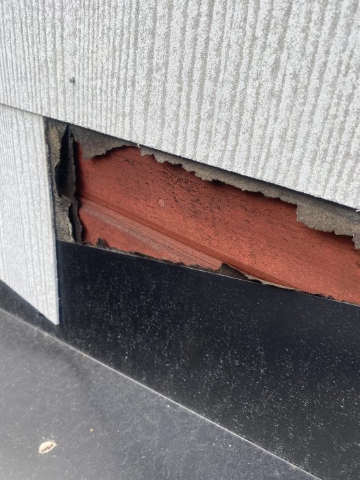 Skadad yttre vägg med ett synligt hål där fasadbeklädnaden har brutits av, exponerande röd isolering undertill.
