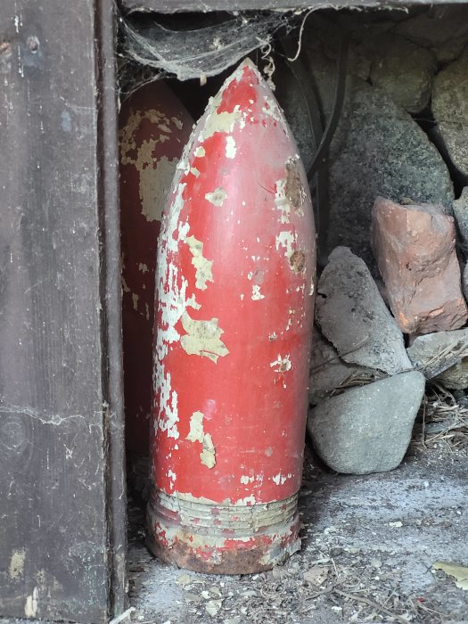 Rödfärgad gammal bomb med avflagad färg står gömd bakom stenar och timmer.