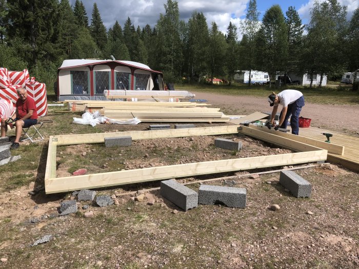 Byggarbetsplats med person som arbetar på golvreglar och isolering, med camping och skog i bakgrunden.