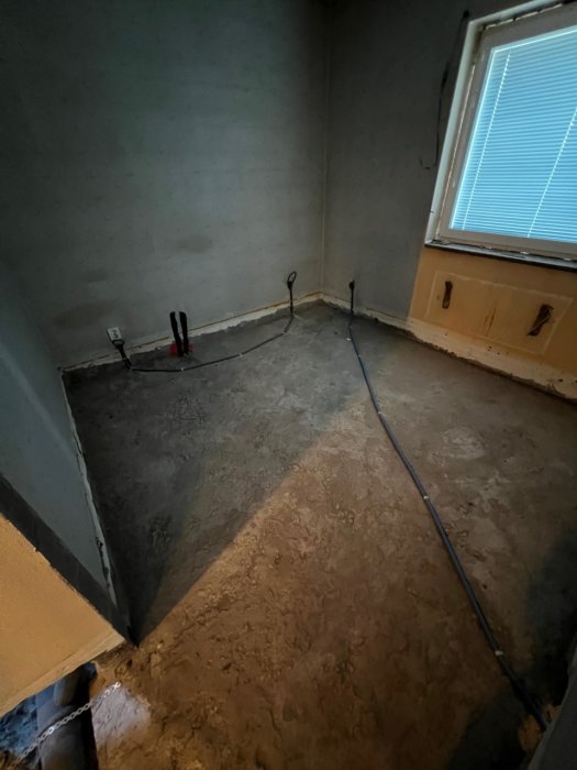 Rått betonggolv i ett rum med synliga slangar och rör framför en vägg och under ett fönster i dagsljus.