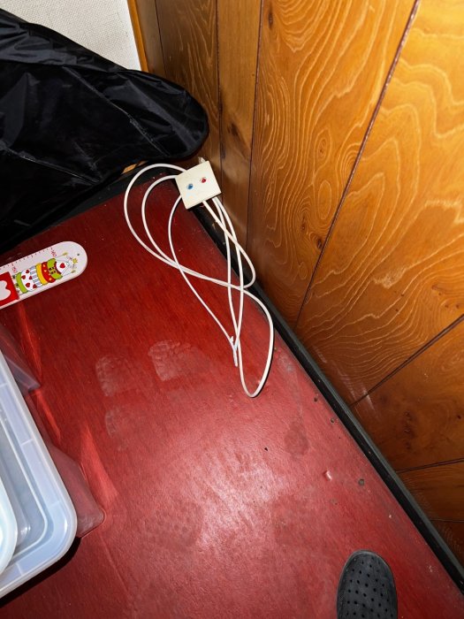En vit kontakt med flera utstickande kablar vid en trähörna intill ett rött golv och en svart plastpåse.