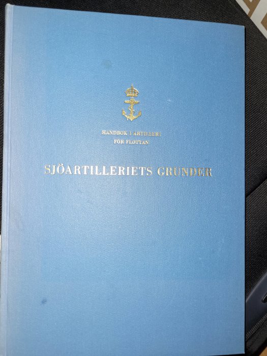 Blå bok med titeln "Sjöartilleriets Grunder" och texten "Handbok i Artilleri för Flottan".