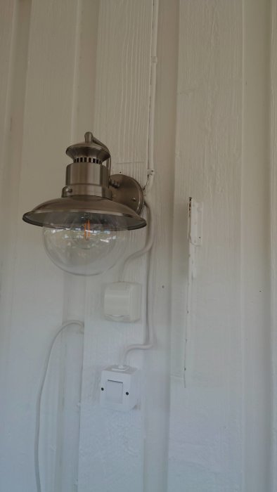 Industriell vägglampa monterad på vit trätak med synliga kablar och strömbrytare.