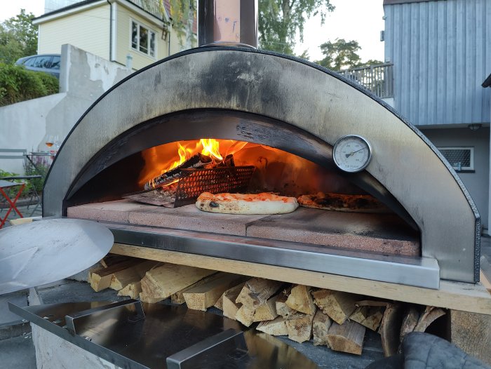 Utomhuspizzaugn med två pizzor som gräddas inuti och ved staplad undertill.