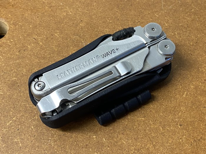 Ett hopfällt Leatherman Wave+ multiverktyg placerat i ett svart, horisontellt 3D-printat fodral på en brun arbetsyta.