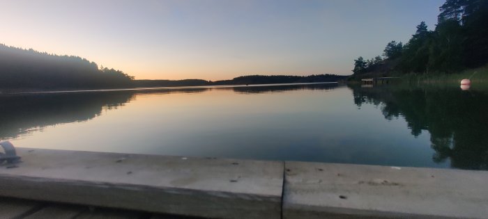 Skymningsvy över en lugn sjö från en brygga, med skogsiluett mot en färggradient i himlen.