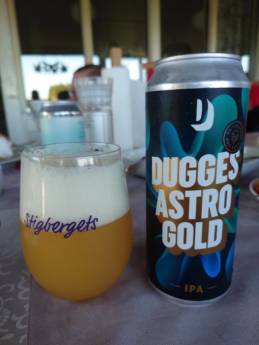 Ölglas med skum på etiketten 'Stigbergets' bredvid en burk Dugges Astro Gold IPA på ett bord.