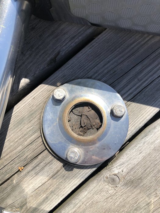 Ett metallfäste med fyra bultar på ett trägolv med ett hål mitt i där röret ska fästas.