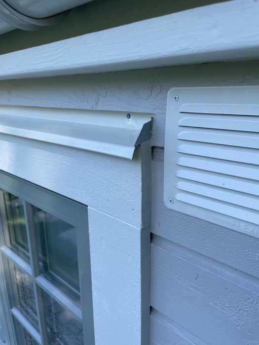 Vitmålad yttervägg med träpanel och ventilkåpa nära ett fönster, detaljbild på husets fasad.