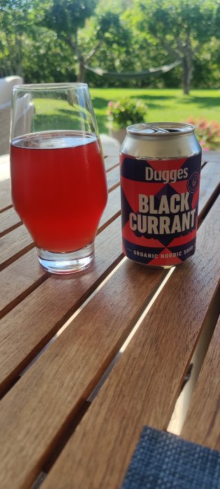 Ett glas röd bärliknande dryck och en burk Dugges Black Current suröl på ett träbord utomhus.