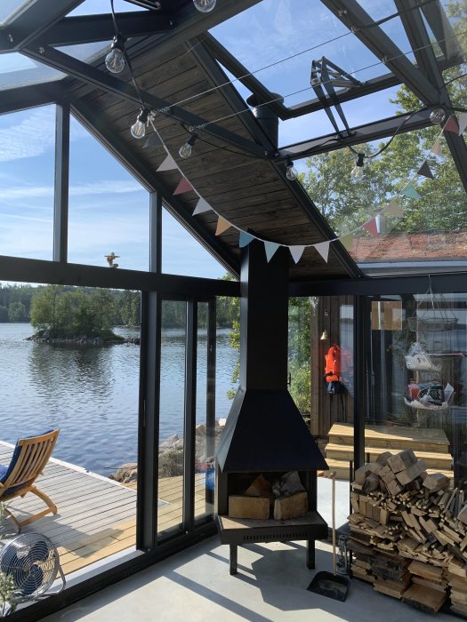 Kamin i glasveranda med ersatt takfönster med råspont och ytpapp, vedtrave bredvid och utsikt över sjö.