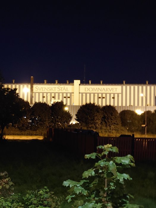 Kvällsbild av industribyggnaden med texten "SVENSKT STÅL DOMNARVET" upplyst på fasaden.