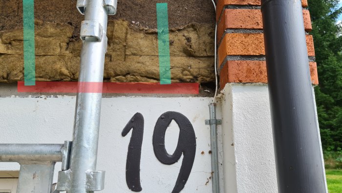 Del av en husvägg med synlig isolering och läkt, vid sidan av siffra 19 och ett stålrör.