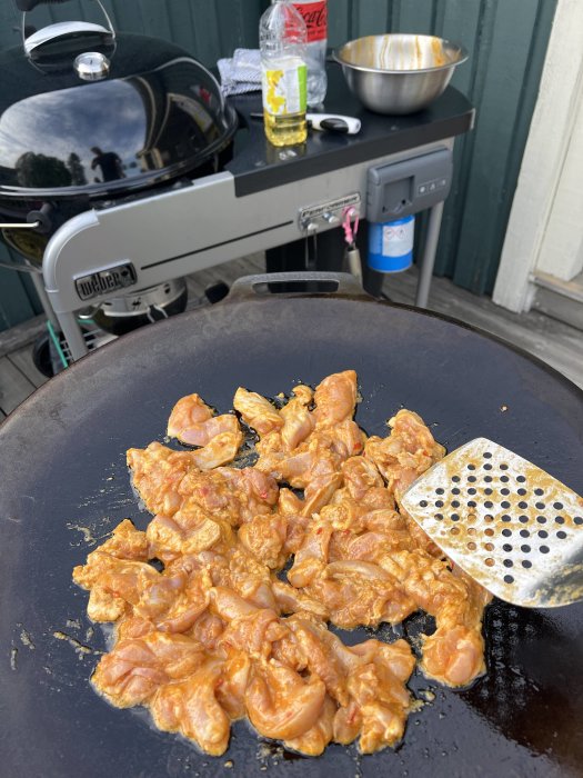 Marinerad kyckling som steks på en grillplatta med grillen och köksredskap i bakgrunden