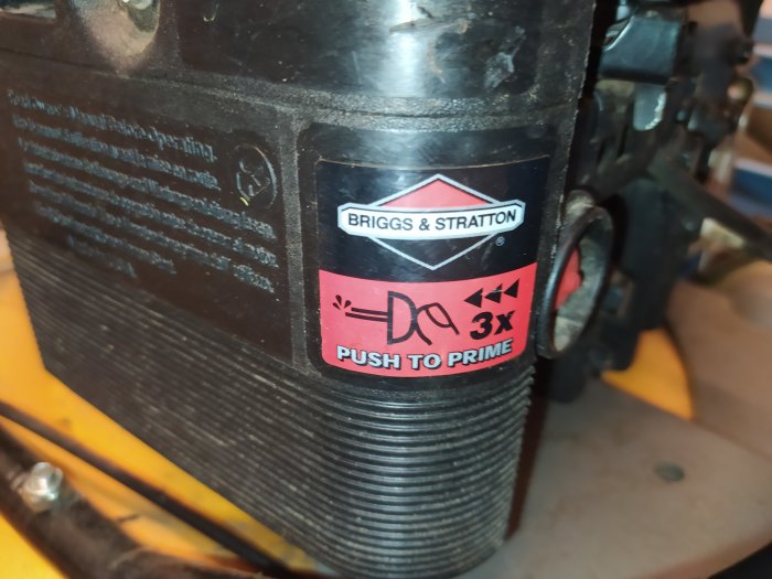 Briggs & Stratton motor med instruktionsetikett som visar "PUSH TO PRIME" på en gul underlag.